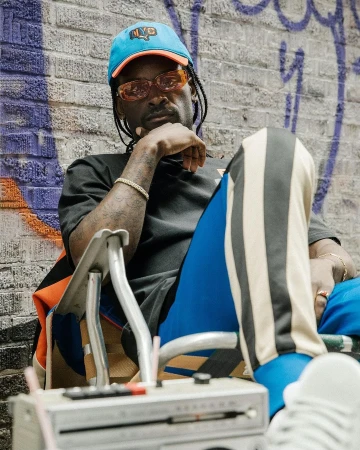 Black man wearing tinted orange sunglasses, blue cap, and striped athletic pants seated outdoors before a brick wall.