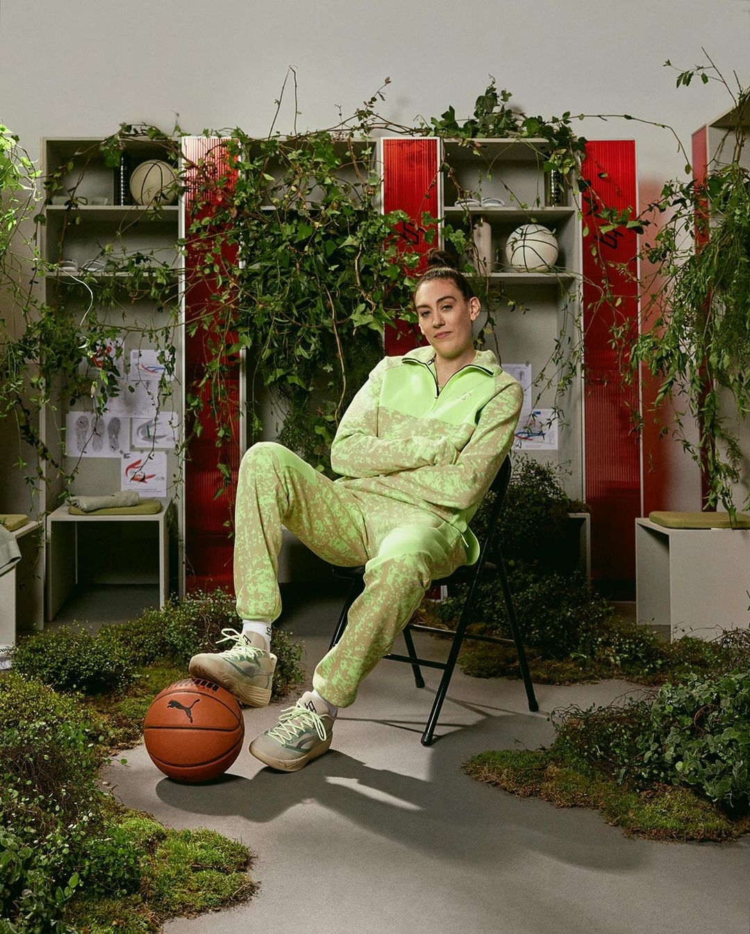 Adult female seated with legs crossed, wearing patterned lime-green tracksuit, basketball at feet, surrounded by overgrown greenery.