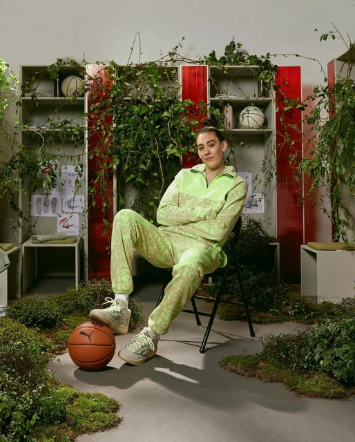 Adult female seated with legs crossed, wearing patterned lime-green tracksuit, basketball at feet, surrounded by overgrown greenery.