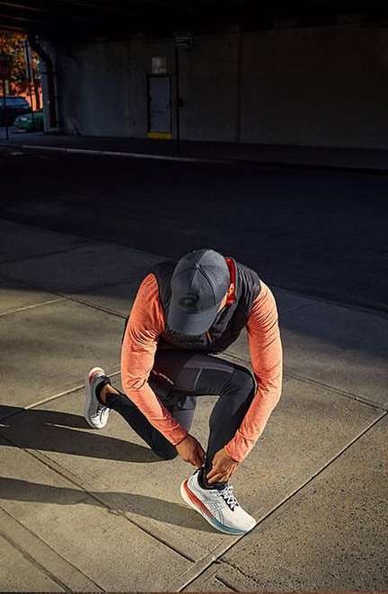 A man kneels on a sidewalk, tying his athletic shoe in an urban setting, with buildings visible in the background and shadows.
