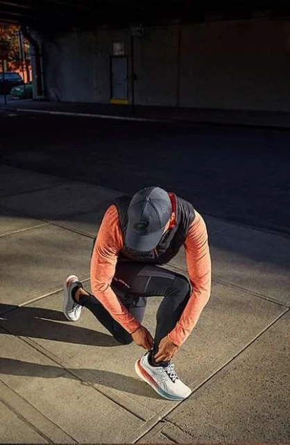 A man kneels on a sidewalk, tying his athletic shoe in an urban setting, with buildings visible in the background and shadows.
