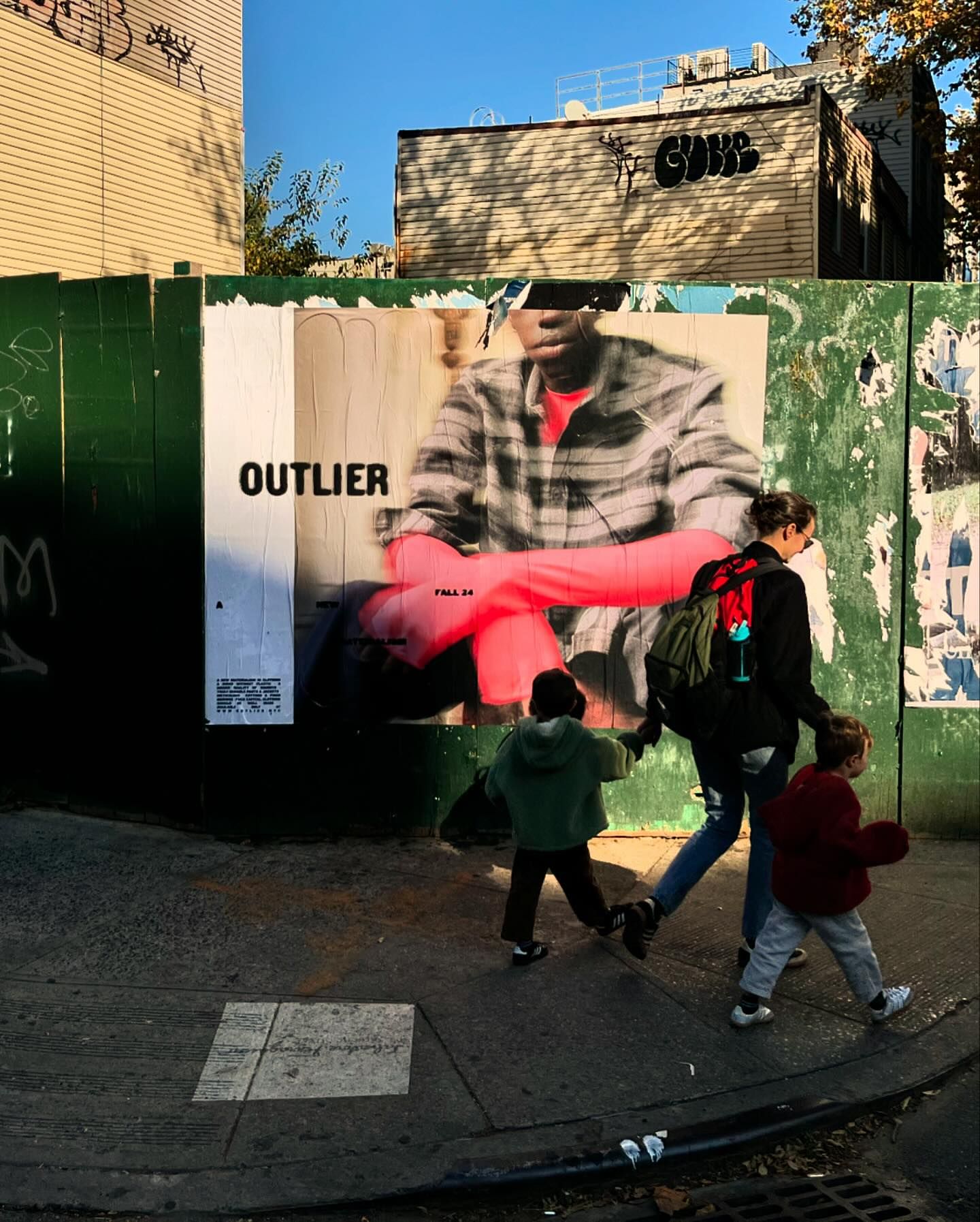 Urban scene with a fashion poster on a green wall, featuring a model in a flannel shirt, and children walking on a sidewalk with their mother.