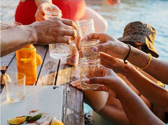 People toasting with clear drinks in mason jars at an outdoor gathering on a wooden deck with a swimming pool in the background.