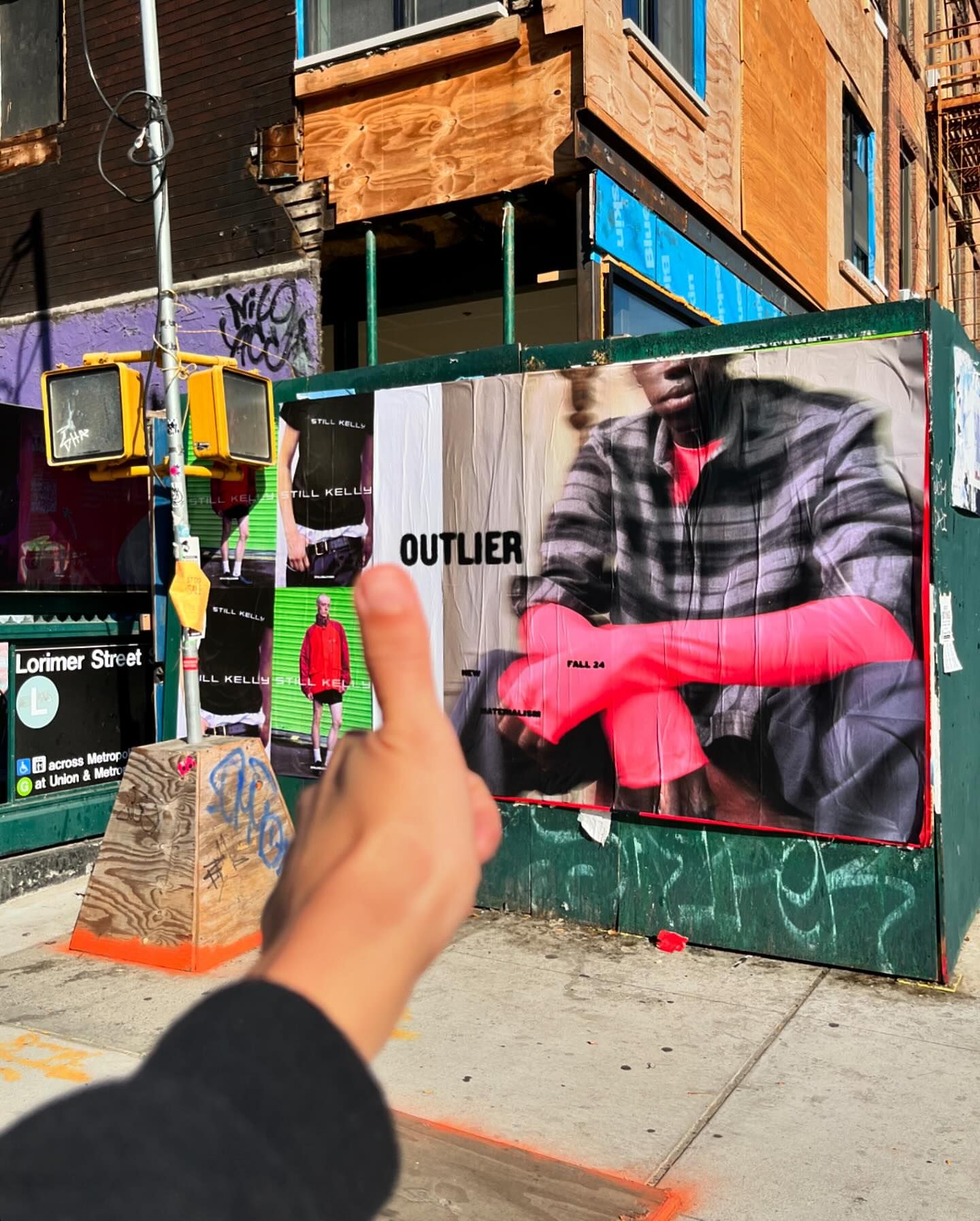 Outdoor shot of a hand gesturing in front of a fashion advertisement featuring a person in plaid and a subway station entrance.