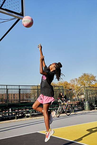 Young woman in pink shorts jumps to make a basket on an outdoor basketball court under a clear sky on a sunny day.