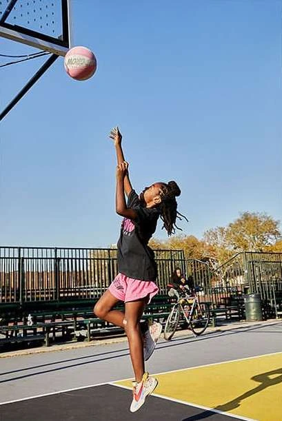 Young woman in pink shorts jumps to make a basket on an outdoor basketball court under a clear sky on a sunny day.