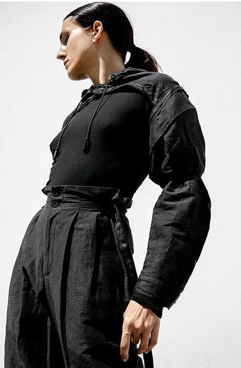 A young adult woman poses indoors in an all-black fashion ensemble with a hooded top, high-waisted pants against a bright white background.