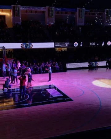 Basketball game in a stadium with a mascot and people on the court, spectators in the stands, and bright pink lighting.