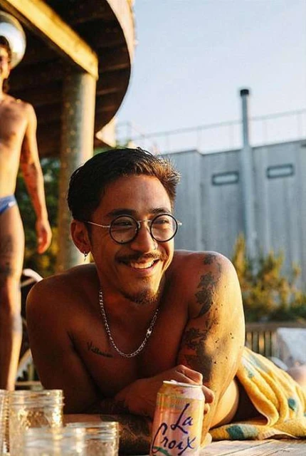 Smiling man with glasses lounges near La Croix cans and glass jars in an outdoor setting with a partial view of another person.
