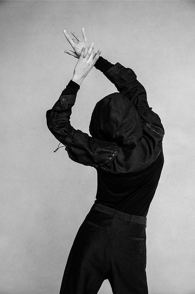 Monochrome studio shot of a person in dark clothing with arms raised, hands posed, and a hooded head against a neutral backdrop.