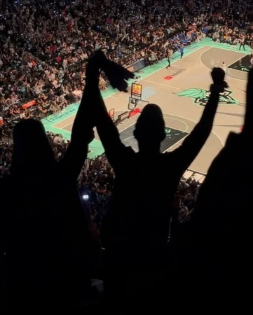 Crowd cheers at indoor basketball game with silhouette of two people in foreground raising arms at sporting event in stadium setting.