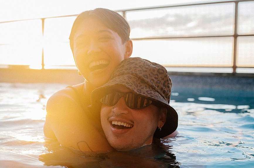 Two smiling adults, one with a bucket hat and sunglasses, embrace in a sunlit pool, creating a warm and joyful scene.