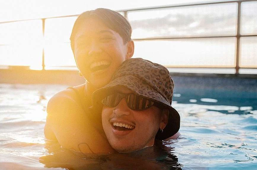 Two smiling adults, one with a bucket hat and sunglasses, embrace in a sunlit pool, creating a warm and joyful scene.