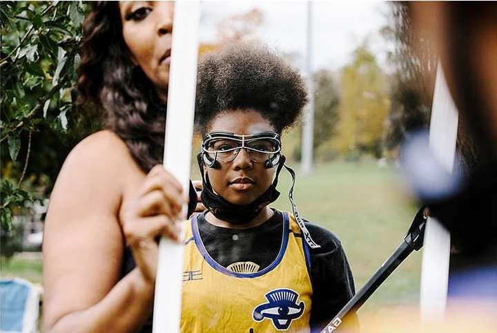 Portrait of a young athlete wearing protective eyewear and sports uniform, with another adult in the background, outdoors in daytime.