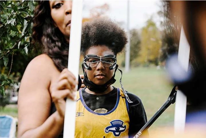 Portrait of a young athlete wearing protective eyewear and sports uniform, with another adult in the background, outdoors in daytime.