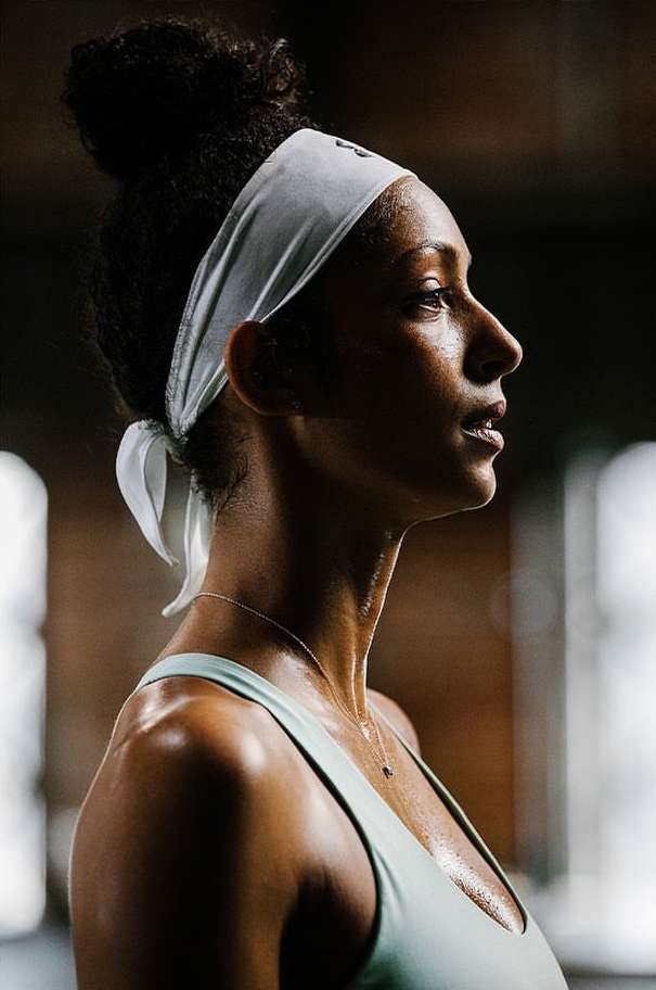 Close-up profile of a sweaty adult woman with dark hair in a bun, wearing a headband and sports top, in a fitness setting.