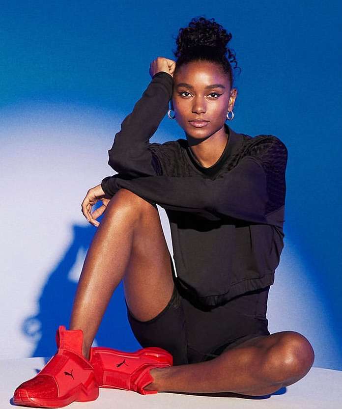 Woman in black athletic wear poses sitting with red Puma sneakers against a split blue and white background in a studio setting.