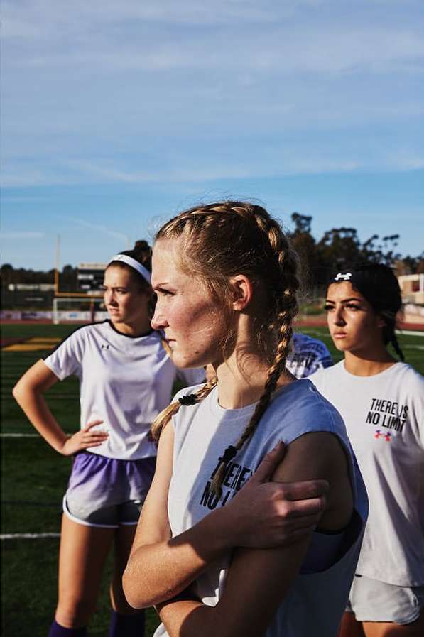 Three female athletes in sports attire stand on a soccer field under a blue sky, facing forward. Focus on the nearest blonde athlete.