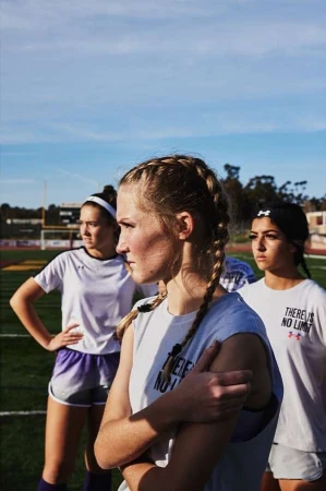 Three female athletes in sports attire stand on a soccer field under a blue sky, facing forward. Focus on the nearest blonde athlete.