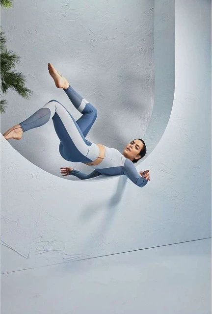 Athletic woman in blue and white activewear strikes dynamic pose mid-air against textured white studio backdrop with green foliage accent.