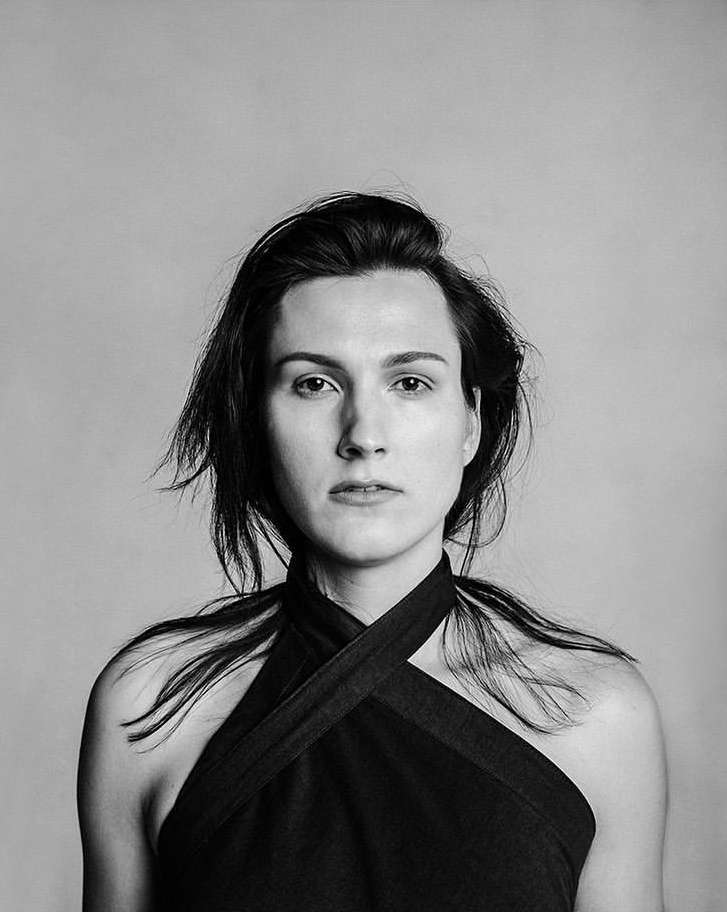 Monochromatic portrait of a woman with dark hair, wearing a halter top, gazing directly at the camera in a minimalist studio setting.