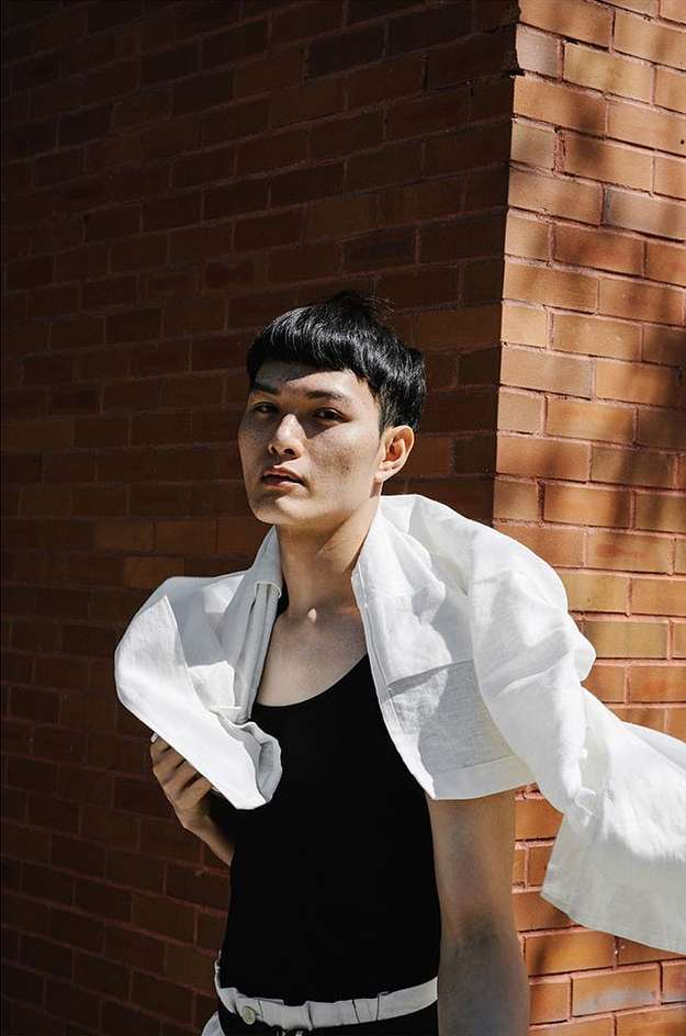 A person in a black shirt and white jacket standing in front of a brick wall in natural light in an urban environment.