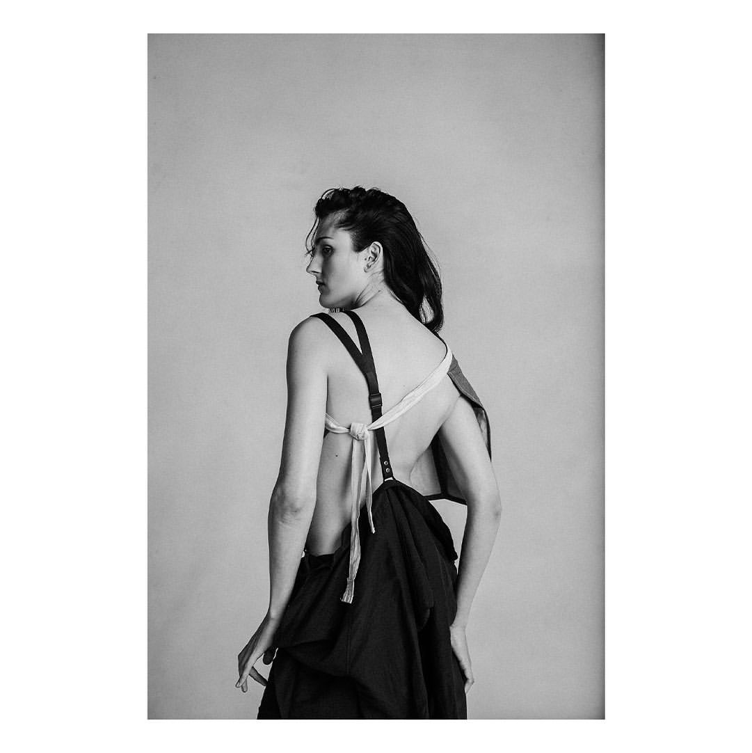 Back view of a woman with dark hair wearing black avant-garde clothing, posing in a studio setting against a neutral background.