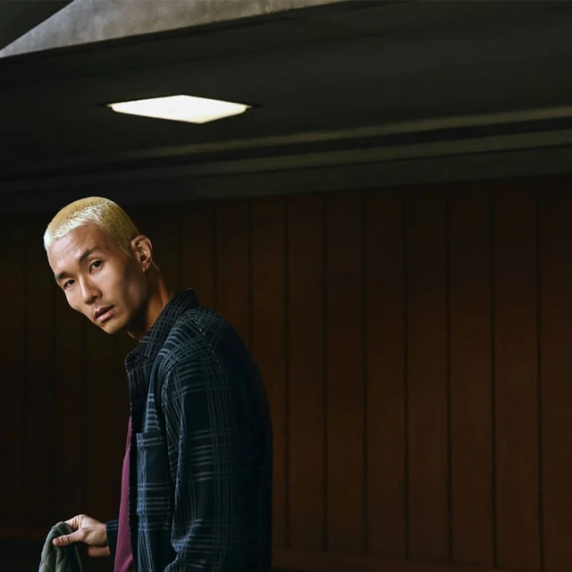 A person with blond hair, wearing a flannel shirt, looking at the camera while standing in an urban indoor environment with concrete walls.