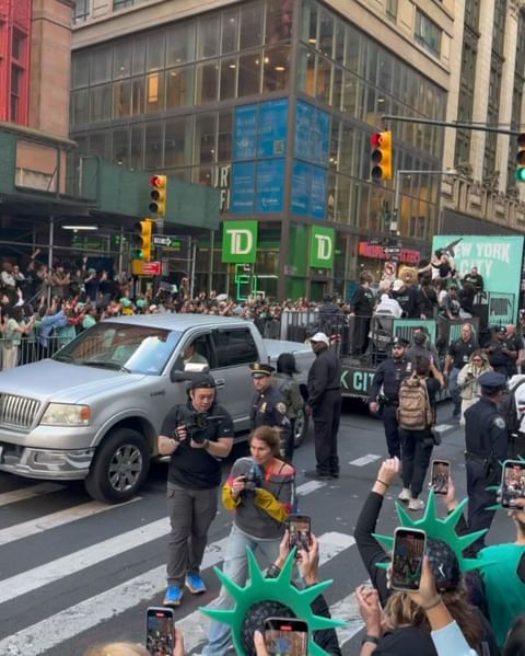 Outdoor shot capturing the Thanksgiving Day parade with crowds, floats, security, and a silver pick-up truck in an urban setting.