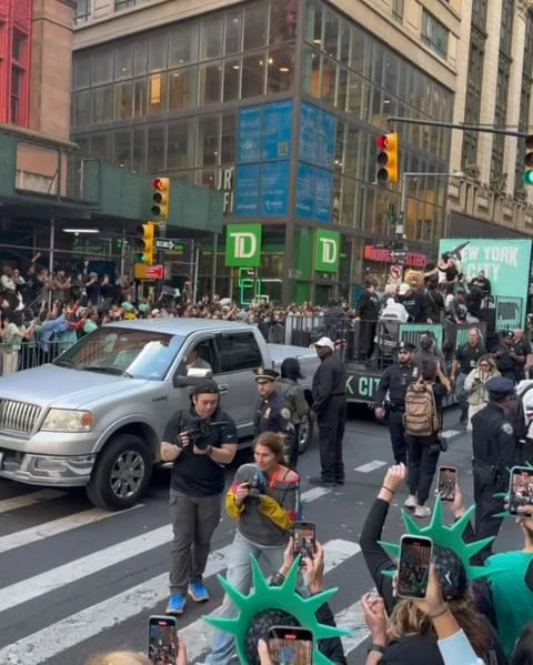 Outdoor shot capturing the Thanksgiving Day parade with crowds, floats, security, and a silver pick-up truck in an urban setting.