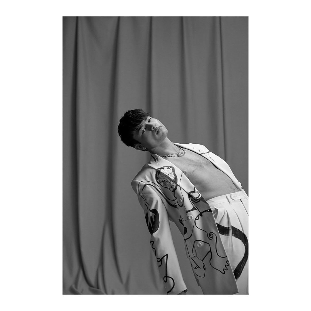 Black and white portrait of a young adult man in an open blazer with graphic line art and white pants, set against a plain curtain backdrop.