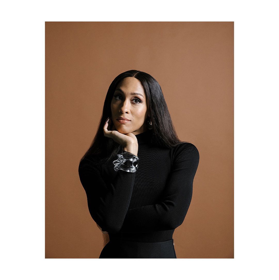 Portrait of a woman with long dark hair, wearing a black turtleneck and a large silver bracelet, posing against a brown backdrop.
