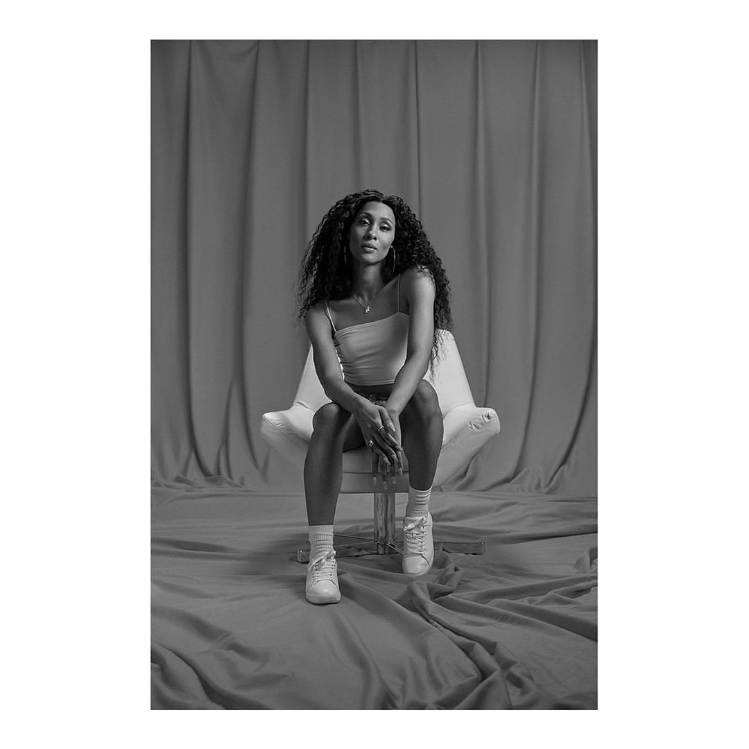 Black and white studio portrait of an adult female seated in a chair with long braided hair, wearing a top, skirt, socks and tennis shoes.