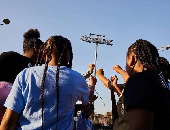 Group of young African-descent people standing outdoors on a sunny day, raising their fists together in unity and solidarity.