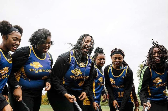 A group of six young Black female athletes, wearing matching athletic uniforms, smiling and standing together on a grassy field outdoors.