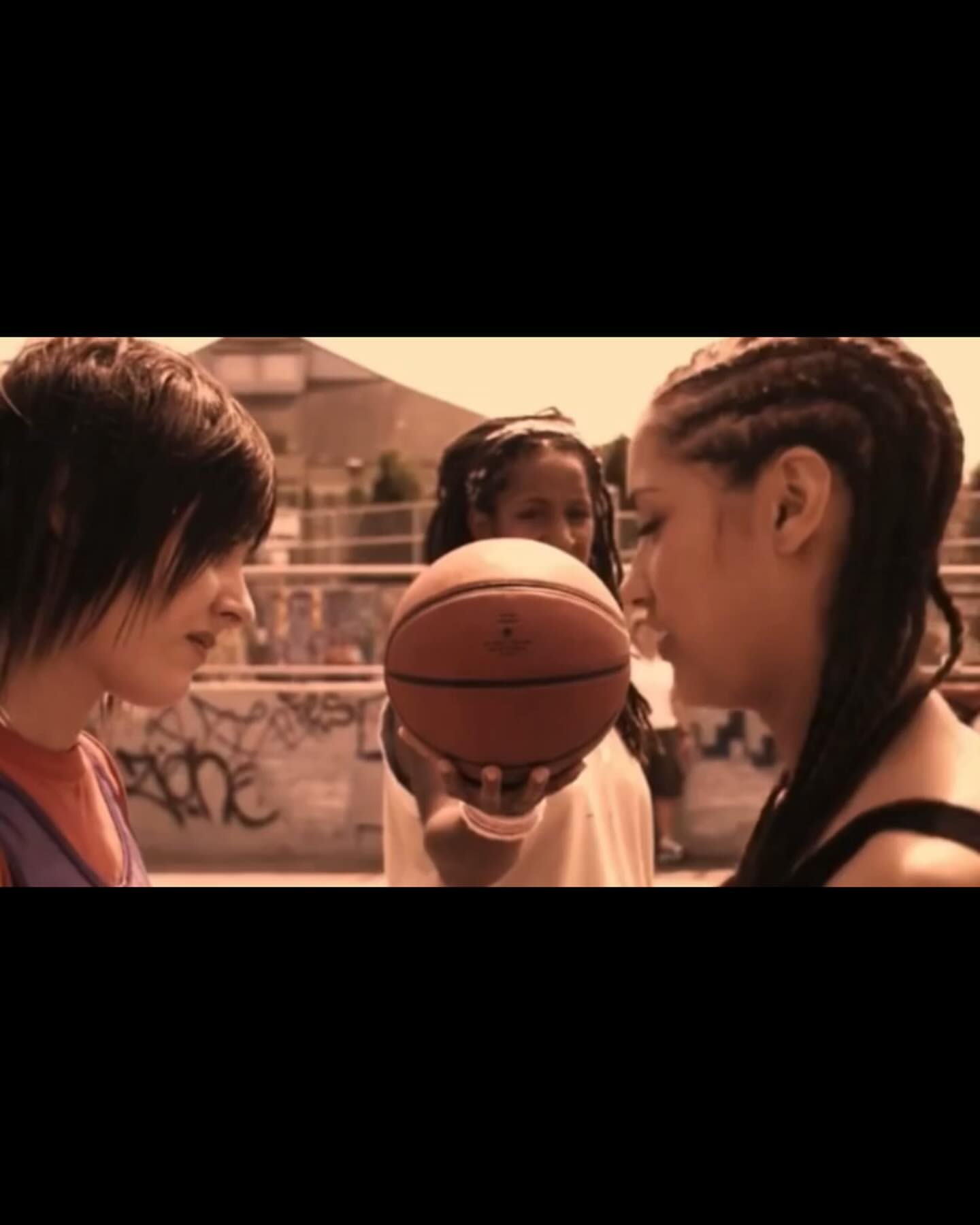 Three young women in casual clothing face each other with a basketball on an outdoor urban court, with graffiti-covered walls visible.
