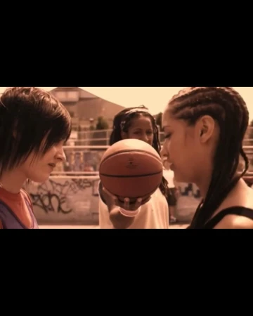 Three young women in casual clothing face each other with a basketball on an outdoor urban court, with graffiti-covered walls visible.