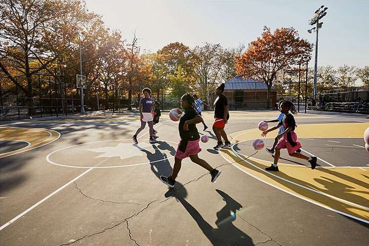 Girls play basketball on an outdoor court surrounded by trees. Children dribble basketballs in an urban park on a sunny day.