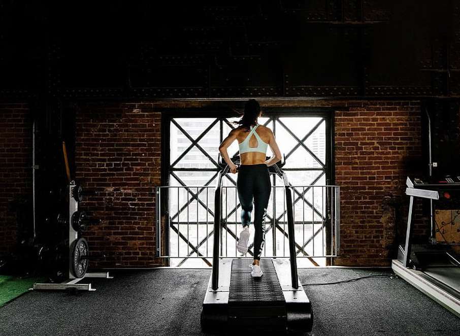 Woman running on a treadmill in a gym with a brick wall, viewed from behind, wearing athletic attire. Fitness and exercise concept.