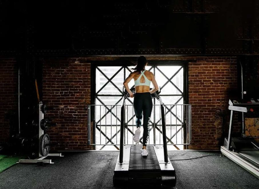 Woman running on a treadmill in a gym with a brick wall, viewed from behind, wearing athletic attire. Fitness and exercise concept.