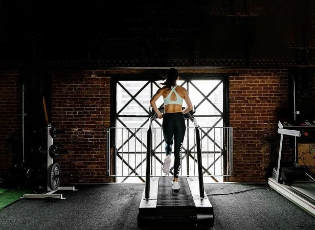 Woman running on a treadmill in a gym with a brick wall, viewed from behind, wearing athletic attire. Fitness and exercise concept.