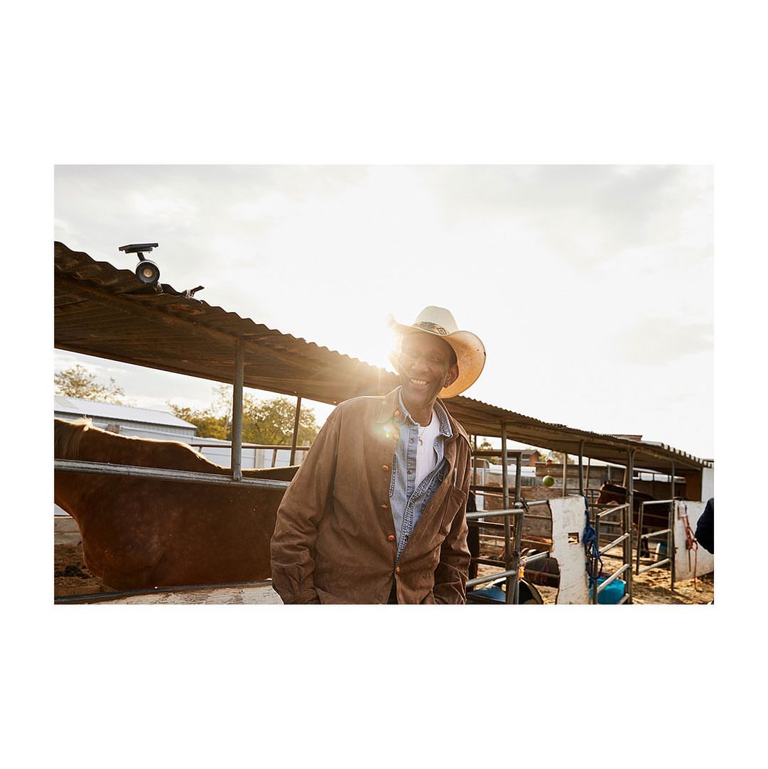 Smiling African-American cowboy in straw hat and brown jacket stands outside stable with horses on a sunny day at eye-level angle.