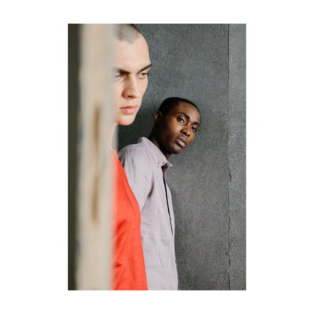 Two young men in a portrait shot, one in red, the other in a lavender shirt against a gray wall, captured in soft natural light.