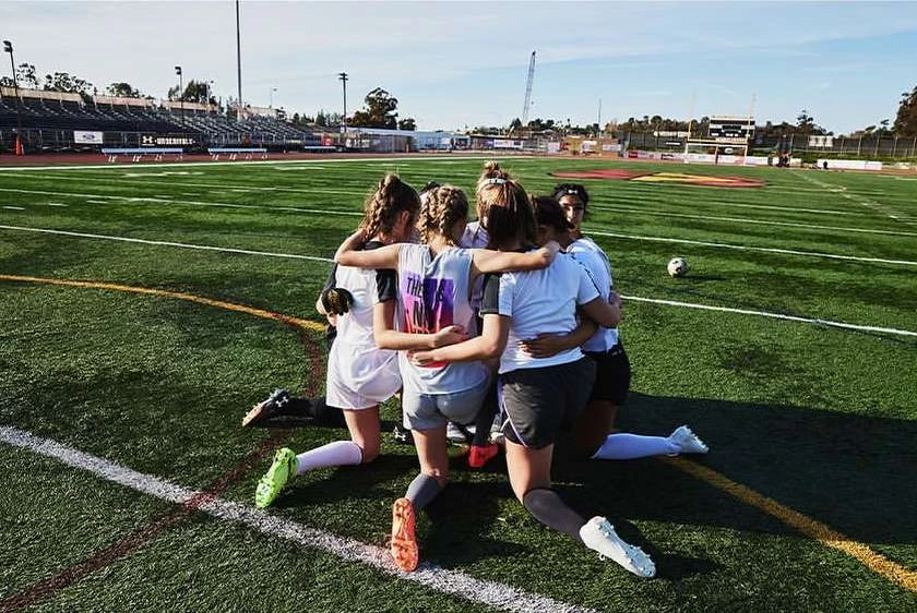 Female soccer team huddles in celebration on a green sports field with stadium seating and a blue sky backdrop. Team spirit and sportsmanship.