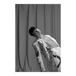Monochrome portrait of a young man wearing a patterned blazer and trousers against a draped backdrop, photographed by Laurel Golio.