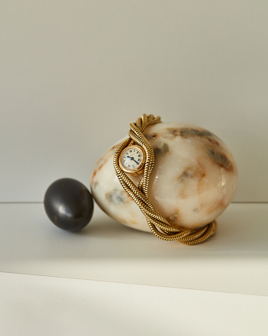 Gold vintage wristwatch with chain bracelet is displayed on marble stone next to black stone on a white surface in a brightly lit studio.