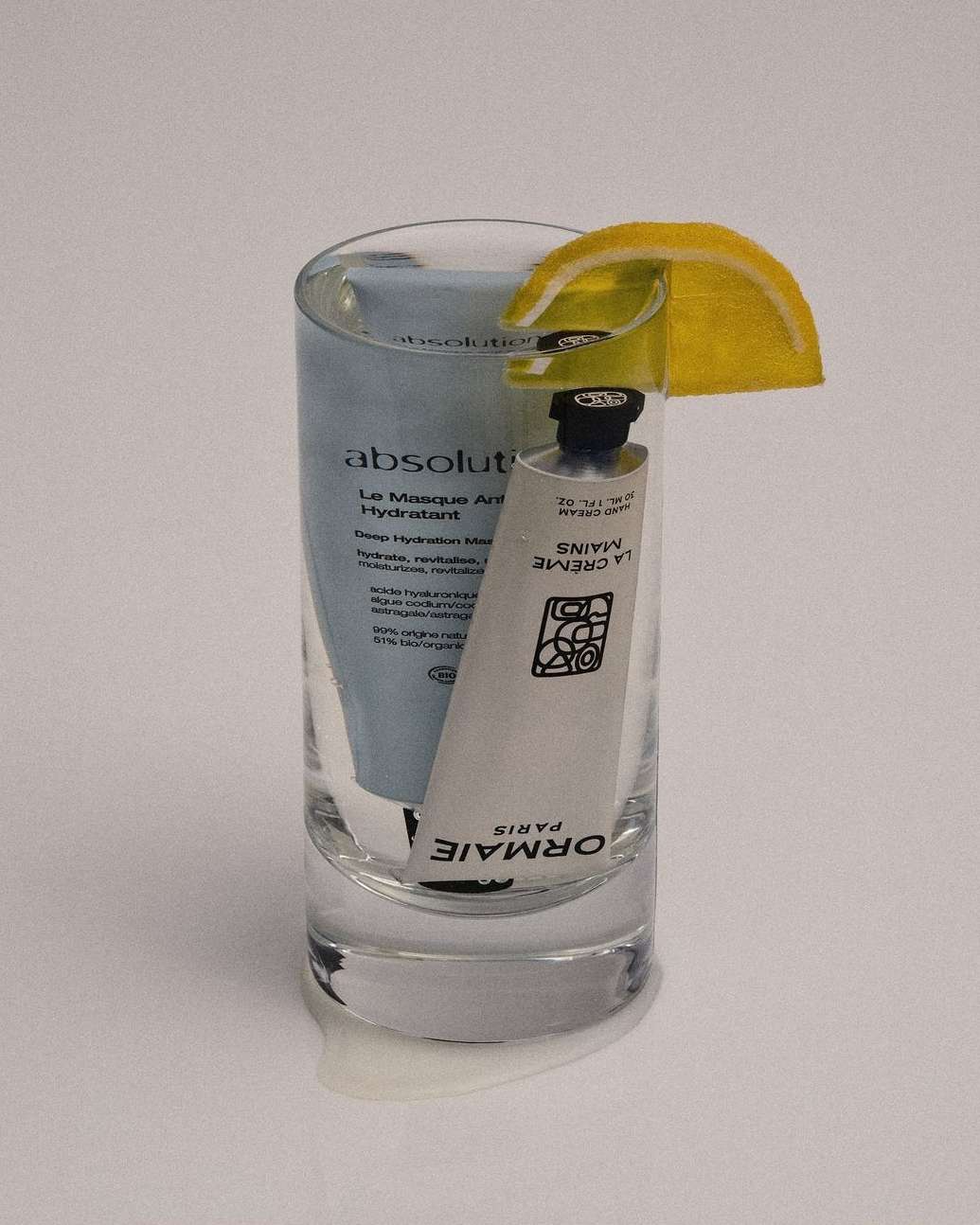 Cosmetic product tubes are arranged in a glass with lemon garnish, evoking a cocktail, set against a soft white background.
