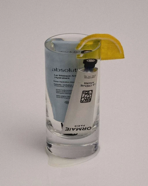 Cosmetic product tubes are arranged in a glass with lemon garnish, evoking a cocktail, set against a soft white background.