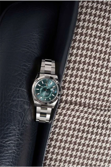 Wristwatch with silver band and green face displayed on a blue leather surface and a houndstooth patterned fabric background.