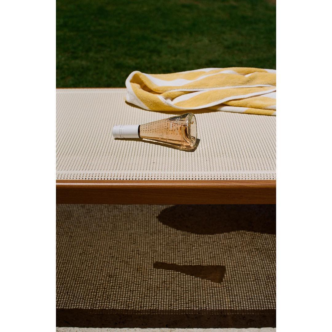 A bottle of rosé wine and a yellow striped beach towel are on a cream colored lounger outdoors on a sunny day.